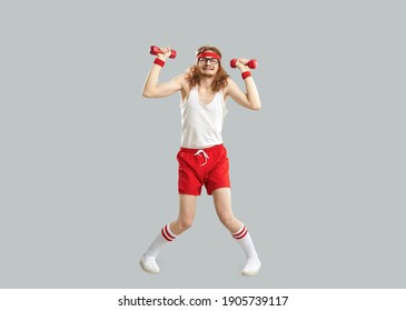 Full body funny tired weak thin skinny man in glasses, retro gym headband, white tank top and red shorts holding heavy fitness dumbbells and doing sports workout exercise isolated on gray background