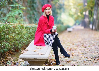 Full Body Of Glad Female In Elegant Beret And Coat, With Paper Cup Of Takeaway Hot Drink, Looking At Camera While Sitting On Bench In Park