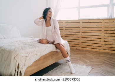 Full Body Of Graceful Young Hispanic Female In Nightwear And Socks Smiling And Looking Away While Touching Dark Hair Sitting On Comfortable Bed With Knitted Plaid At Home