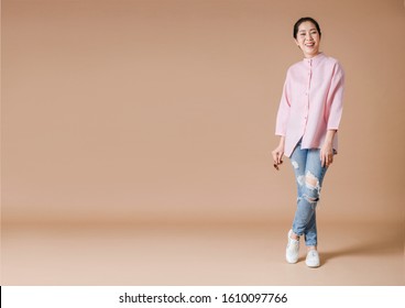 Full Body Of An Happiness Asian Woman Wearing Pink Shirt, Holding Sunglasses In Right Hand And Standing On Beige Background.