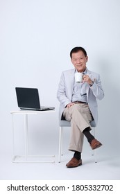 Full Body Of An Happy Asian Old Man In Light Blue Suit And Beige Pants Drink Coffe And Working With Laptop In Studio White Background.