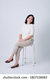 Full Body Of An Happy Asian Old Woman In White Shirt And Beige Pants Sitting In Studio White Background.