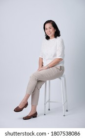 Full Body Of An Happy Asian Old Woman In White Shirt And Beige Pants Sitting In Studio White Background.