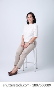 Full Body Of An Happy Asian Old Woman In White Shirt And Beige Pants Sitting In Studio White Background.