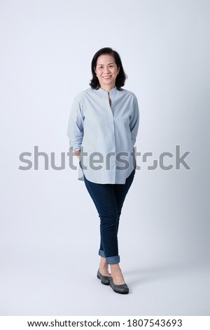 Full body of an happy asian old woman in light blue shirt and dark blue pants standing in studio white background.