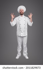Cuerpo completo de feliz cocinero barbudo con uniforme de chef blanco y gorro demostrando gestos correctos contra fondo gris