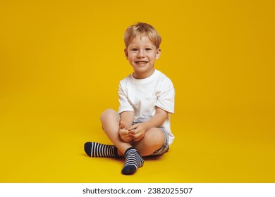 Full body of happy blonde child boy wearing casual clothes, smiling and looking at camera while sitting crossed legged on floor, isolated over yellow background. - Powered by Shutterstock