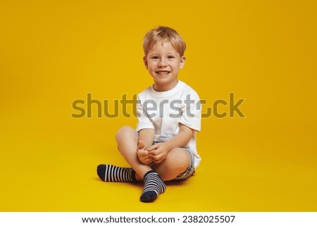 Full body of happy blonde child boy wearing casual clothes, smiling and looking at camera while sitting crossed legged on floor, isolated over yellow background.