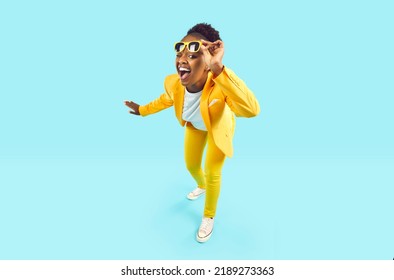Full body happy cheerful Afro American woman wearing stylish yellow suit and trendy glasses standing isolated on blue background, looking at camera, winking her eye and smiling. Party, fashion