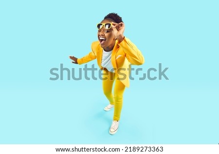 Full body happy cheerful Afro American woman wearing stylish yellow suit and trendy glasses standing isolated on blue background, looking at camera, winking her eye and smiling. Party, fashion Foto stock © 