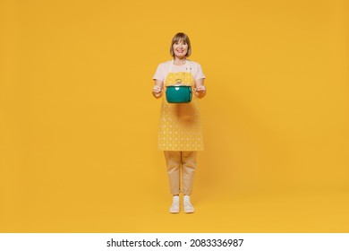 Full body happy elderly housekeeper housewife woman 50s in orange apron hold green saucepan with soup look camera isolated plain on yellow background studio portrait People household lifestyle concept