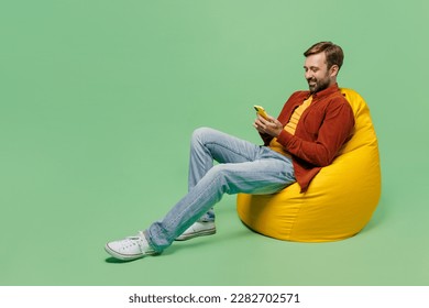 Full body happy elderly man 40s years old he wears casual clothes red shirt t-shirt sit in bag chair hold in hand use mobile cell phone isolated on plain pastel light green background studio portrait - Powered by Shutterstock