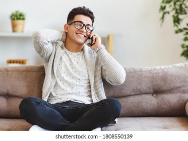 Full Body Happy Ethnic Man In Casual Clothes And Glasses Rubbing Neck And Looking Away With Smile While Sitting Cross Legged On Couch And Answering Phone Call At Home
