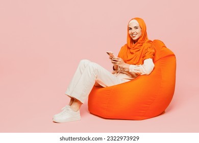 Full body happy fun young arabian asian muslim woman wears orange abaya hijab sit in bag chair hold use mobile cell phone isolated on plain pink background. Uae middle eastern islam religious concept - Powered by Shutterstock