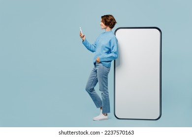 Full body happy side view young woman wear knitted sweater near big huge blank screen mobile cell phone with workspace mockup area use smartphone isolated on plain pastel light blue cyan background - Shutterstock ID 2237570163