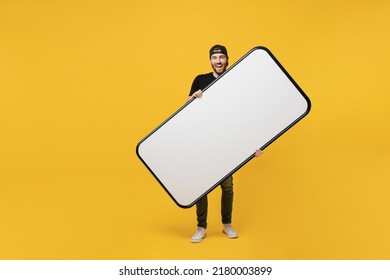 Full Body Happy Smiling Fun Cool Young Tattooed Man He 20s Wears Black T-shirt Cap Holding Big Huge Blank Screen Mobile Cell Phone Workspace Mockup Area Copy Space Isolated On Plain Yellow Background