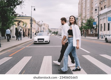 Full body of happy young couple in casual clothing holding hands while crossing road on crosswalk in Moscow town during daytime