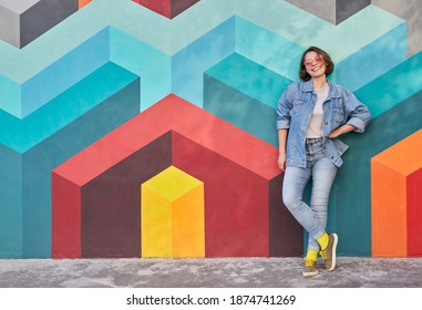 Full body happy young female hipster in casual clothes smiling and leaning on wall with geometric ornament while resting on city street
