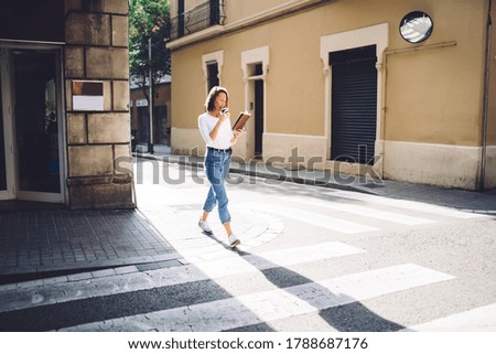 Full body of happy young female in casual clothes drinking hot coffee while walking on street and reading interesting book