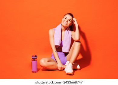 Full body happy young fitness trainer instructor woman sportsman wear top shorts purple clothes in home gym sit drink water relax rest isolated on plain orange background. Workout sport fit concept