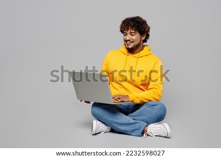 Full body happy young IT Indian man 20s he wearing casual yellow hoody sitting using holding working on laptop pc computer isolated on plain grey background studio portrait. People lifestyle portrait