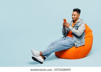 Full body happy young man of African American ethnicity wear denim jacket orange t-shirt sit in bag chair hold in hand use mobile cell phone isolated on plain pastel light blue cyan background studio - Powered by Shutterstock