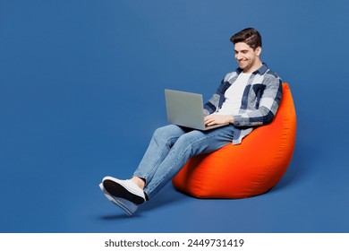 Full body happy young IT man he wear shirt white t-shirt casual clothes sit in bag chair hold use work on laptop pc computer isolated on plain blue cyan background studio portrait. Lifestyle concept