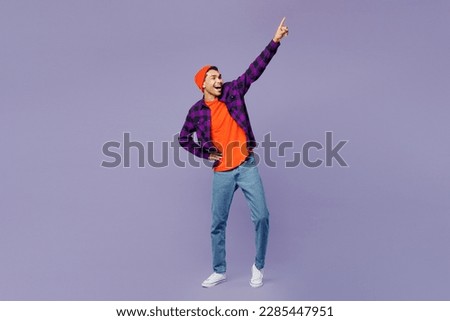 Full body happy young man of African American ethnicity wear casual shirt orange hat stand akimbo raise up hand point finger up isolated on plain pastel light purple color background studio portrait