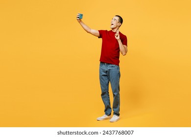 Full body happy young middle eastern man he wear red t-shirt casual clothes doing selfie shot on mobile cell phone post photo on social network show v-sign isolated on plain yellow orange background