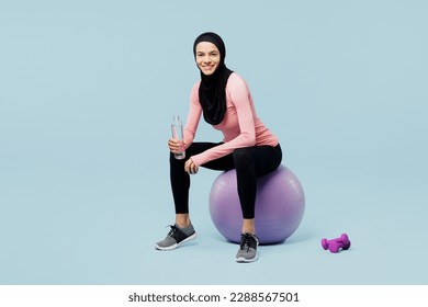 Full body happy young muslim fitness trainer sporty woman wear pink abaya hijab spend time in home gym sit on fitball drink water isolated on plain blue background studio Workout sport fit abs concept - Powered by Shutterstock