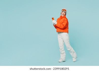 Full body happy young skier woman wear warm padded windbreaker jacket hat ski goggles mask use mobile cell phone travel rest spend weekend winter season in mountains isolated on plain blue background
