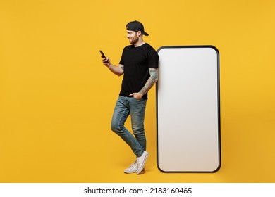 Full Body Happy Young Tattooed Man He Wears Black T-shirt Cap Near Big Huge Blank Screen Mobile Cell Phone Workspace Mockup Area Use Smartphone Isolated On Plain Yellow Background Tattoo Translate Fun