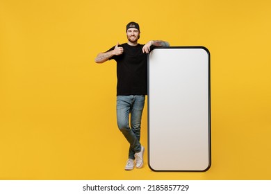 Full Body Happy Young Tattooed Man He Wears Black T-shirt Cap Near Big Huge Blank Screen Mobile Cell Phone Workspace Mockup Area Show Thumb Up Isolated On Plain Yellow Background Tattoo Translate Fun