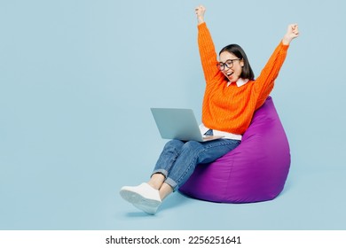 Full body happy young IT woman of Asian ethnicity wear orange sweater glasses sit in bag chair work hold use laptop pc computer do winner gesture isolated on plain pastel light blue cyan background - Powered by Shutterstock