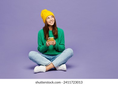 Full body happy young woman wear green sweater yellow hat casual clothes sit hold in hand use mobile cell phone look aside isolated on plain pastel light purple background studio. Lifestyle concept