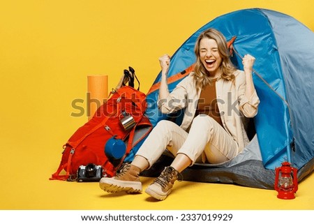 Full body happy young woman sit near bag with stuff tent do winner gesture isolated on plain yellow background. Tourist leads active lifestyle walk on spare time. Hiking trek rest travel trip concept