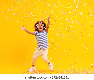 Full body of joyful little black child with Afro hair in stylish clothes laughing and jumping while trying to catch colorful confetti against yellow background