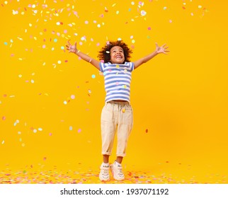 Full body of joyful little black child with Afro hair in stylish clothes laughing and jumping while trying to catch colorful confetti on birthday against yellow background - Shutterstock ID 1937071192
