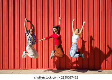 Full Body Of Joyful Multiethnic Female Friends Looking At Camera And Jumping Near Red Wall While Having Fun On Sunny Day