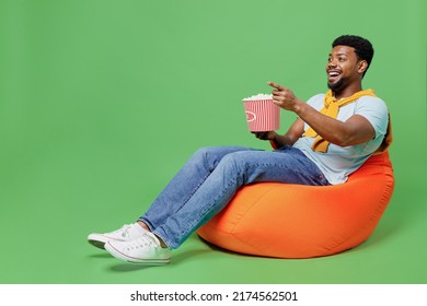 Full Body Laughing Fun Young Man Of African American Ethnicity Wear Blue T-shirt Sit In Bag Chair Hold Popcorn Bucket Point Finger Aside Watch Movie Isolated On Plain Green Background Studio Portrait