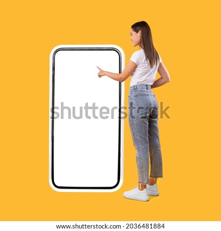 Full Body Length Back View Of Woman Using Big Smartphone With Blank White Screen And Touching Display Panel With Finger, Cheerful Lady Standing On Yellow Background, Ordering Food Delivery, Mock Up Photo stock © 