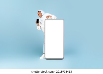 Full Body Length Of Black Muslim Woman Peeking Out, Standing Behind Big Smartphone With White Blank Screen And Holding Cellphone, Presenting New App Or Cool Website, Mockup