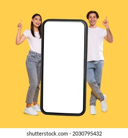 Full Body Length Of Cheerful Couple Standing Next To Big Smartphone With Blank White Screen, Pointing Up, Recommend New Mobile Application On Yellow Studio Background, Mock Up For Design, Ad Template