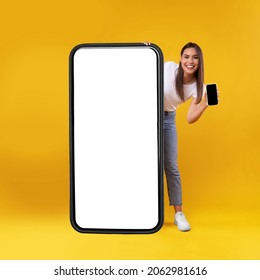 Full Body Length Of Cheerful Woman Peeking Out Standing Behind Big Smartphone With White Blank Screen, Excited Lady Holding Cell Phone Presenting New App Showing Copy Space For Website Design Mock Up
