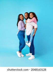 Full body length of funny black family of three people posing on blue studio background, copy space, vertical shot. Cheerful African American girl piggybacking on her daddy, standing together with mom