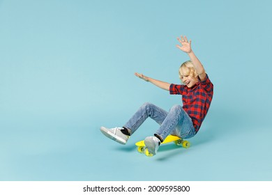 Full body length funny little curly kid boy 13s in basic red checkered shirt sitting riding on skateboard rising hands isolated on blue background children studio portrait. Childhood lifestyle concept