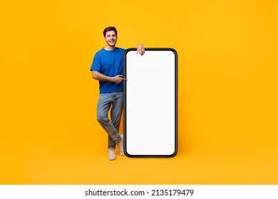Full Body Length Of Happy Casual Guy Pointing Finger At Big Blank Smartphone Display Leaning On Huge White Empty Cell Phone Screen Standing On Yellow Studio Background Mockup. Mobile App Advertisement - Shutterstock ID 2135179479