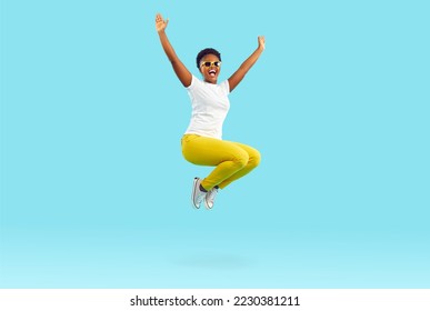 Full body length happy cheerful positive crazy excited young African American woman in white T shirt, yellow pants and sunglasses enjoying weekend or holiday mood and jumping in air on blue background - Powered by Shutterstock