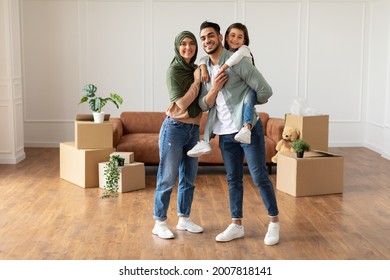 Full Body Length Of Happy Islamic Dad, Mum In Headscarf And Little Girl Posing In New Apartment, Standing In Empty Living Room With Cardboard Boxes. Cheerful Father Carrying His Daughter On Back