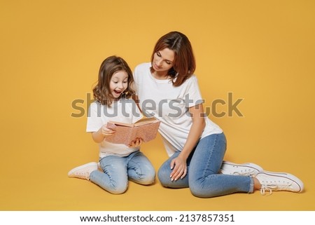 Full body length happy woman in basic white t-shirt sit on floor read book child baby girl 5-6 years old Mom mum little kid daughter isolated on yellow color background studio Mother's Day love family